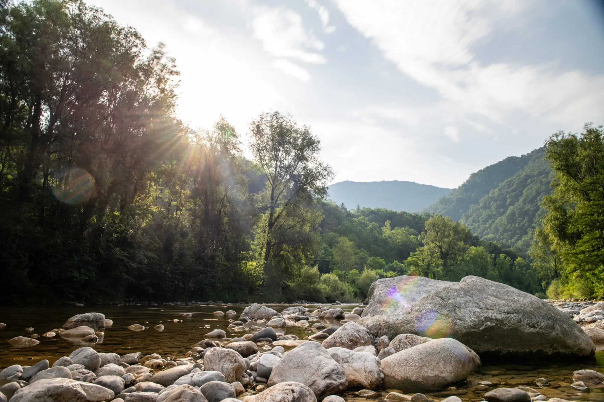 Sun flaring through trees over a rocky river bed with wooded hills behind