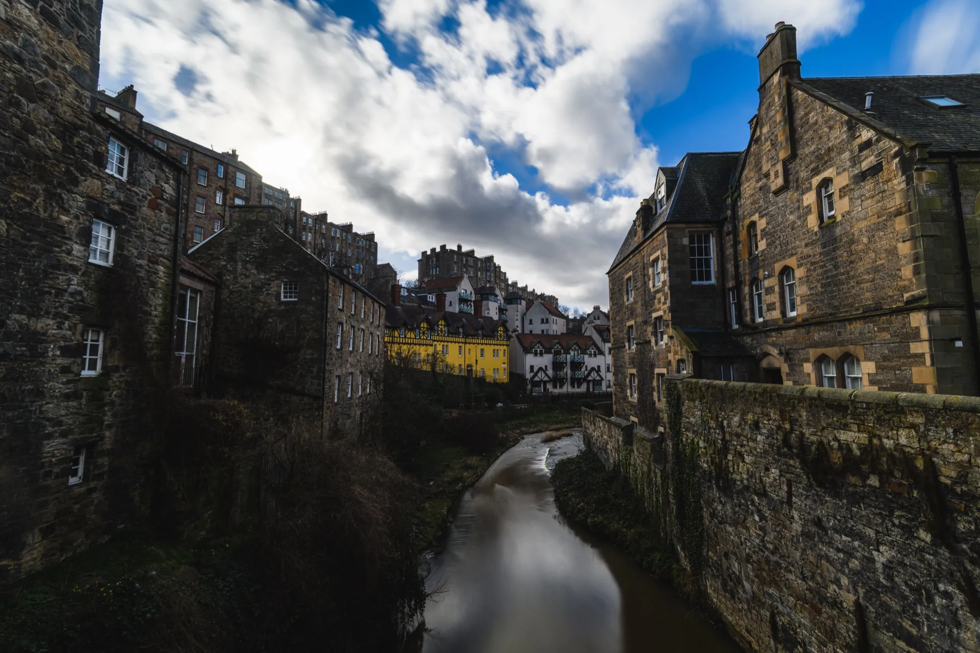 Stone houses and a yellow building along a narrow river in Dean Village, Edinburgh