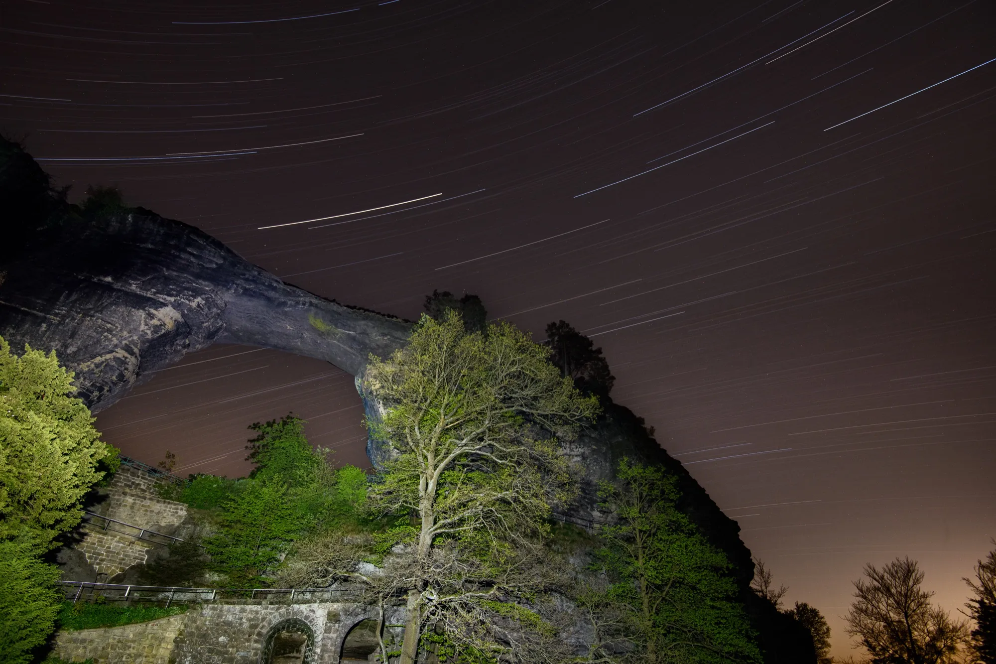 Star trails over a natural rock arch and spring trees at night
