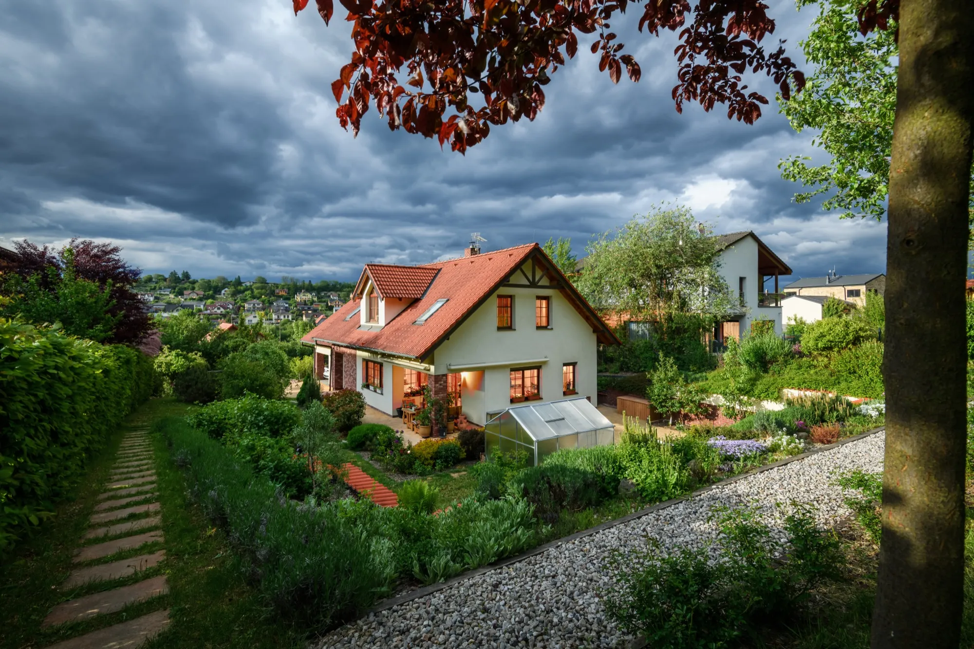 A house with a red roof and lit windows in a garden under a storm sky