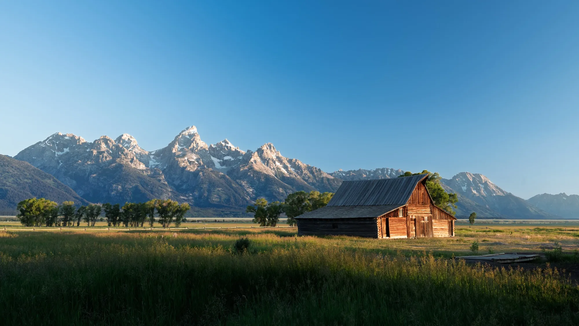 An old wooden barn in a grassy field below the Teton range at sunrise