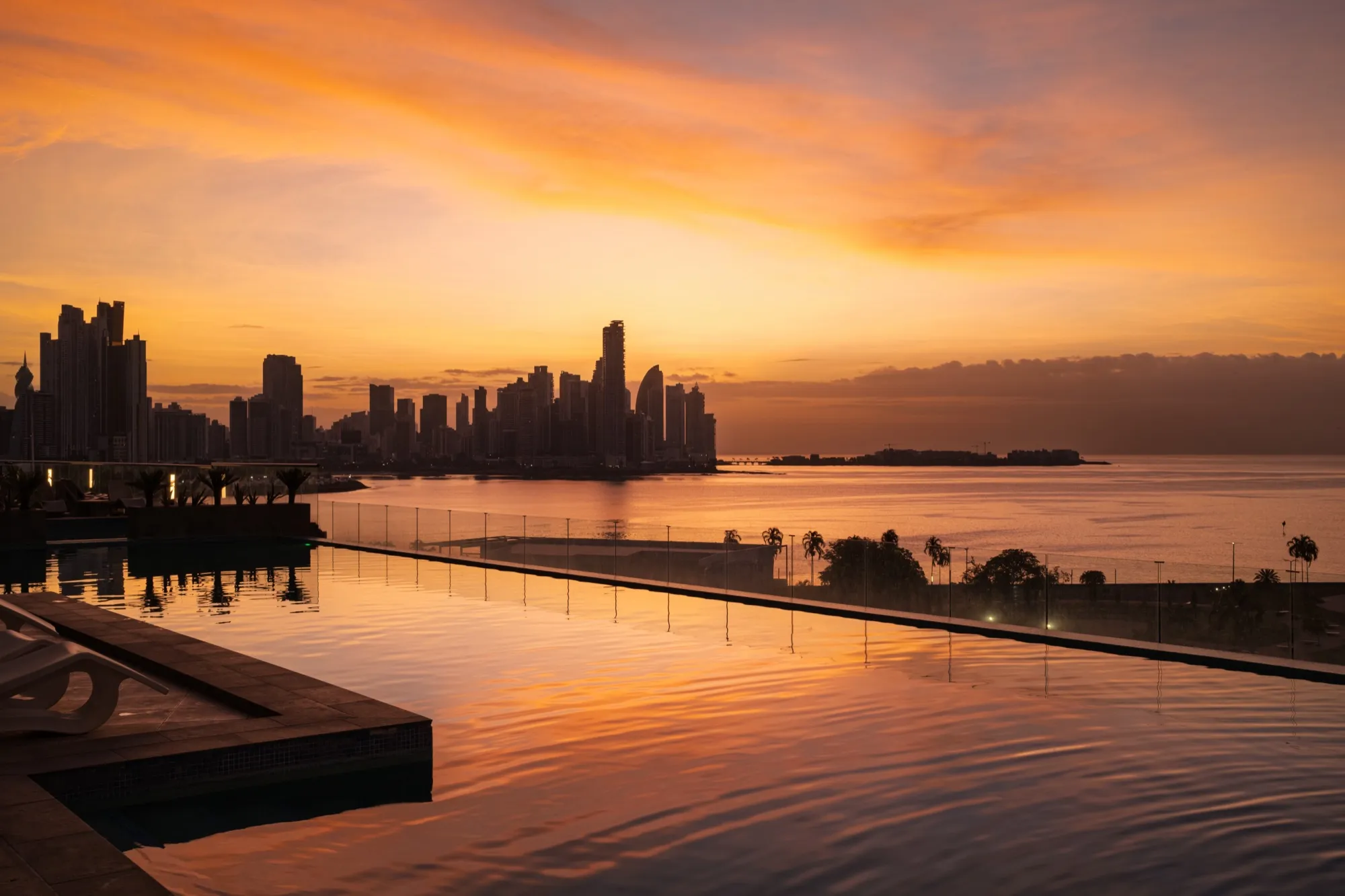 A city skyline across the water at sunset, seen over an infinity pool